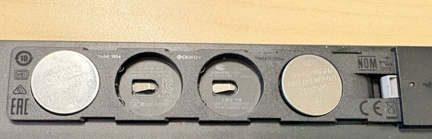 Close-up of the keyboard's underside battery bay: four slots in a row, the shell marked CR2032 with a positive-side-up symbol, cells in slots one and four, slots two and three empty showing the metal contacts at the bottom